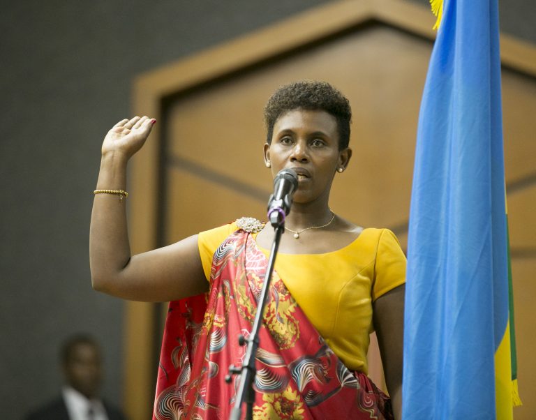 Claudine Uwera svärs in som minister. Hon står bredvid Rwandas flaggs och håller upp sin högra arm. Framför henne står en mikrofon.