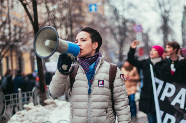 Olga Shevchenko går i ett demonstrationståg. Hon använder en megafon. I bakgrunden syns fler demonstranter.